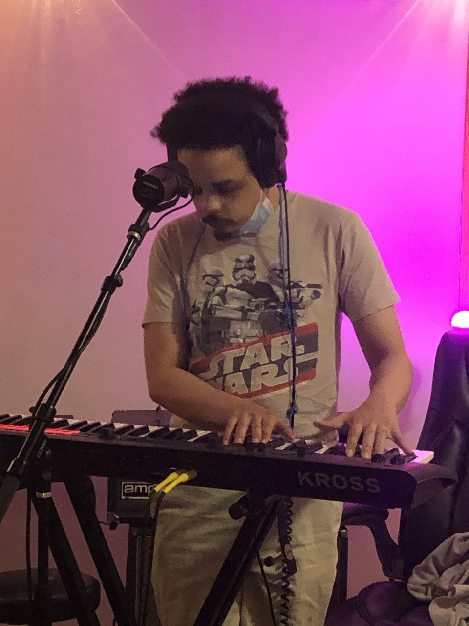 A blind man dressed in a Star Wars T-shirt, depicting storm troopers, plays keyboard with a microphone in front of him and a pink screen in the background. He wears black headphones and a surgical mask pulled down around his chin.