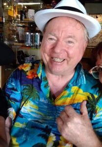 A person smiles for the camera while giving a thumbs up. They are wearing a fedora and Hawaiian style button shirt. 