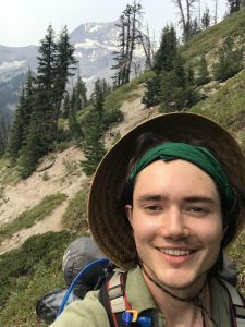 A young man poses for a selfie while on a hiking on a mountain trail. He is wearing a sun hat and button-up shirt and carrying a large utility backpack filled with camping equipment.
