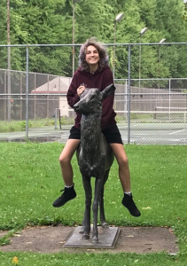 Zoe is riding on an animal statue in a park.  Their feet are off the ground and they look happy.  The animal is carved out of a stone and appears to be a deer.