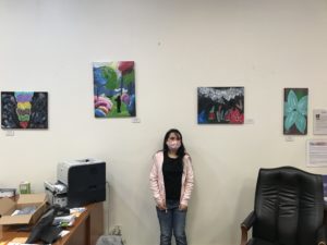 Image of artist Ariana Rangel standing in front of a wall of her paintings at the BBVA Bank.