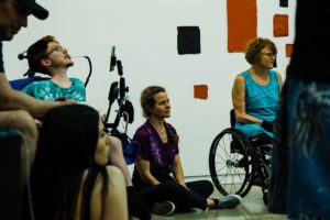 People sitting in a circle in the gallery room with white walls and blue oblong installation made out of different shades and sizes of rectangular and squares. Some people are on the floor, some on their wheelchairs having smile on their faces.