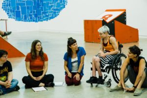 People sitting either on the floor or in their wheelchairs and behind them there is red and black rectangular and squares of different sizes painted on the wall. Everyone is looking off camera but in the same direction.