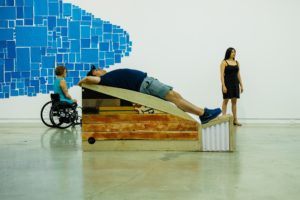 gallery room with blue oblong made of different sized cubes and rectangular on the wall and in the middle of the image there is an arch build from wood where there is person laying on top of it on their back. On the left side of them there is a person in wheelchair and on the right side of them there is a person standing in front of a white wall.