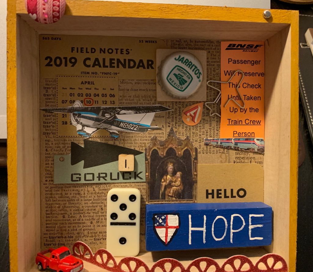A shallow box filled with overlapping found objects such as page from a dictionary, scrabble letter, domino, images of planes, and words such as Hope, Hello, and Groruck.