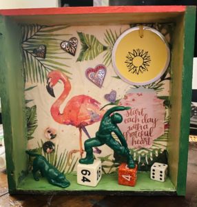 A shallow box filled with overlapping found objects such as die, small green army man and dog in yoga poses, a paper napkin with a flamingo on it, a stamped tag, hearts, and a quote.