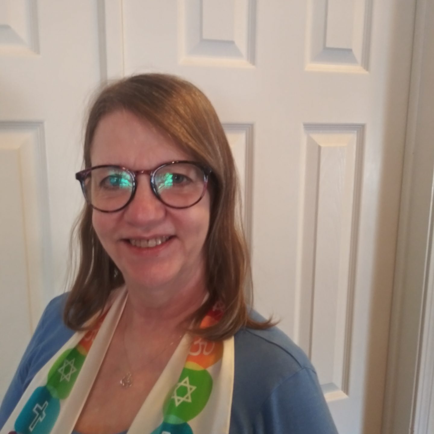 A woman with circular framed glasses and shoulder length brown hair smiles for the camera. She is wearing a scarf with a variety of religious symbols on it.