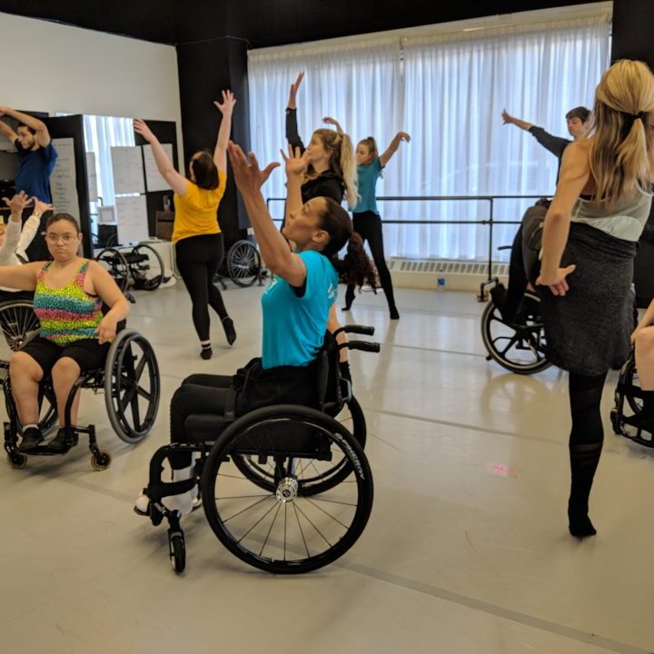 Sit-up and stand-up dancers dancing in a studio that has a big window in the background.