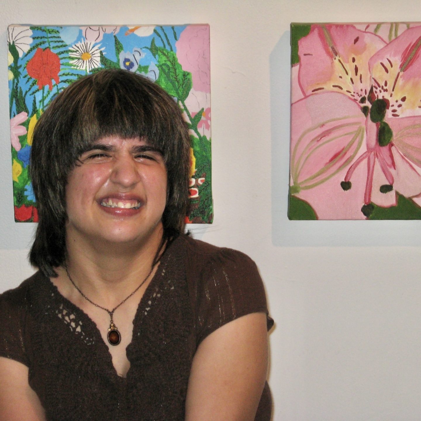 An artist smiles in front of a wall where their artwork is hung.