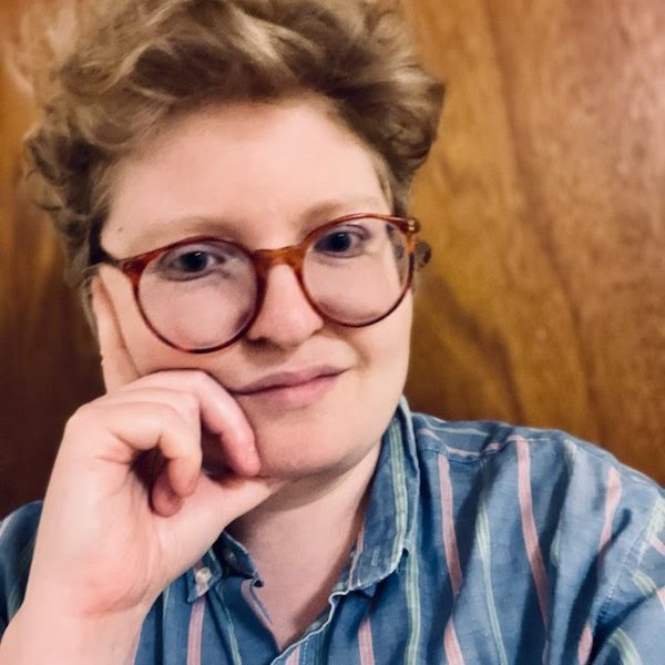 Photo of Joey Gidseg, a white nonbinary person with short blonde curly hair and big red glasses, resting their chin on their hand with a slight smile. Joey is wearing a light blue shirt with pink and white stripes and is in front of a wood paneled background.