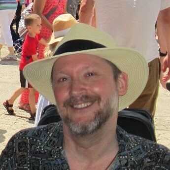 A caucasian man smiles, while seated. He has brown hair and a brown mustache, and a brown and white beard. He is wearing a patterned green, black and white shirt that is unbuttoned at the neck and a pale yellow hat with a wide brim and a black band around it. In the background are multiple people. A young boy with blonde hair, a red shirt and black shorts with a white stripe walks next to a woman in a pink and white dress. There is also another person wearing a white t-shirt and khaki shorts.