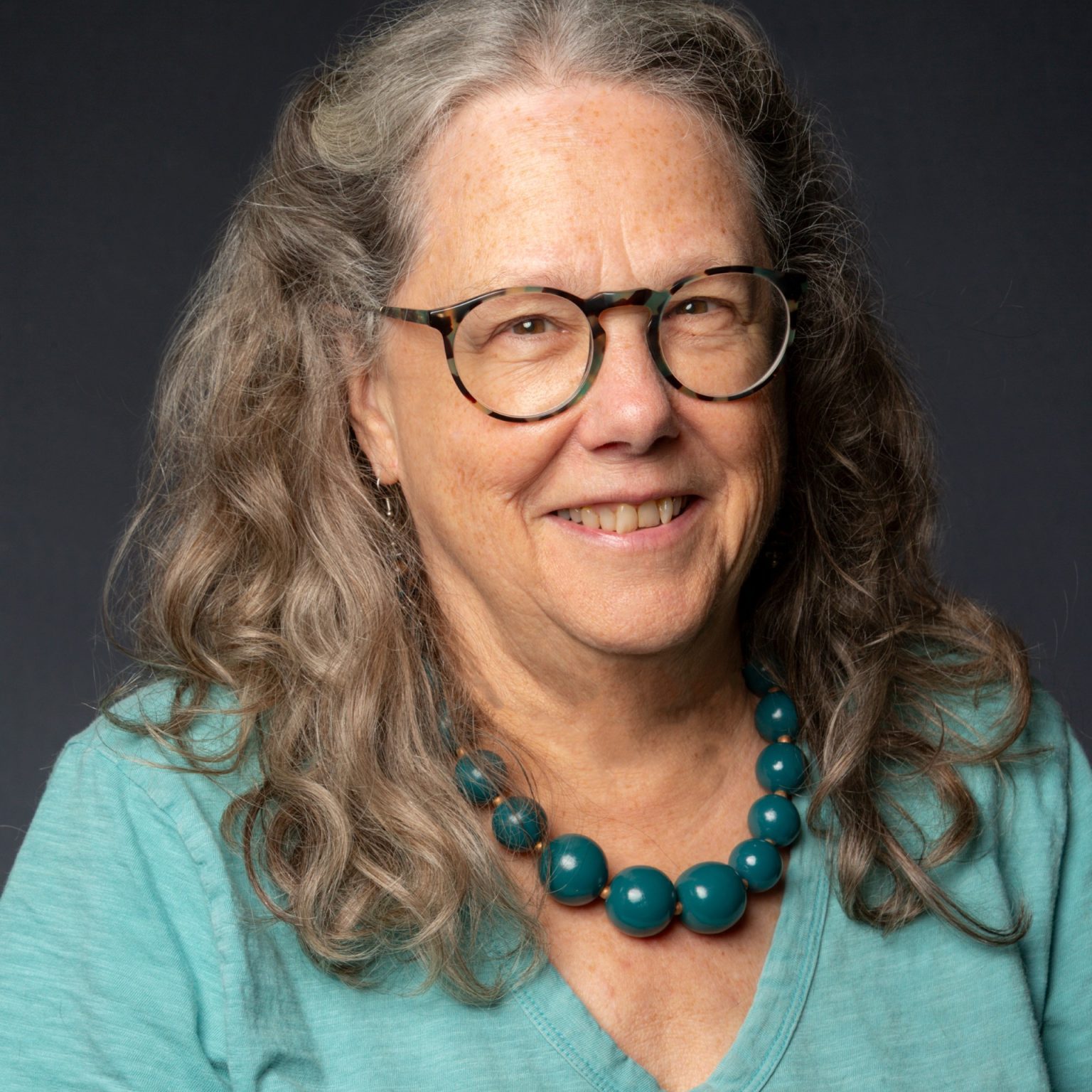 Celia Hughes wearing a light blue top with a large dark blue beaded necklace and black rimmed glasses with grey curly/wavy hair