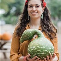 A woman with long brown hair, dangling earrings, and red headband smiles in a pumpkin patch while holding a guard.