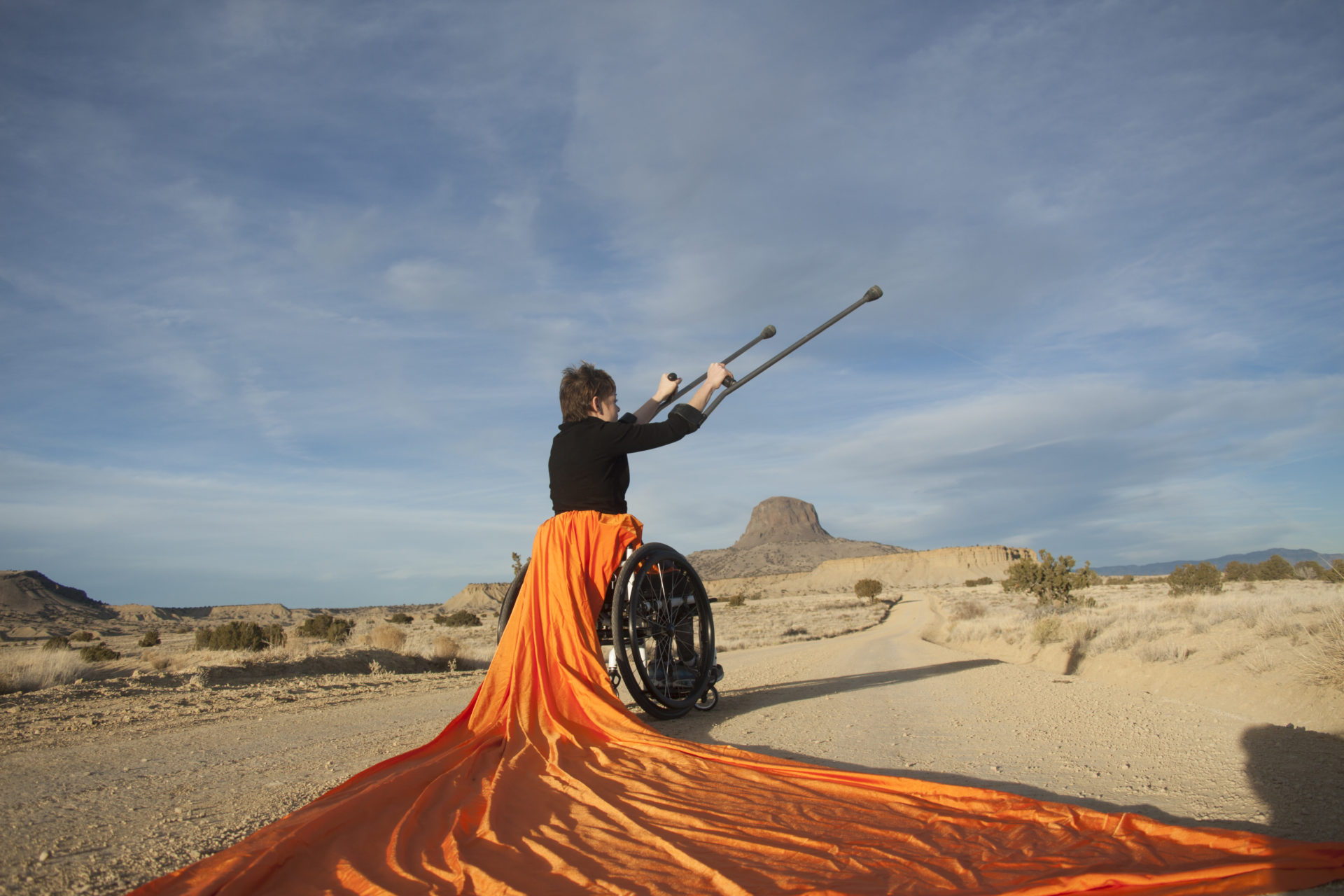 Tanya sitting in her chair on a dirt road and the chair has large orange fabric draped form the seat of it. Tanya is holding her crutches diagonally up towards the blue and white sky. The photo is taken in New Mexico and has desert landscape with small hill on it. Photo by Pat Perret