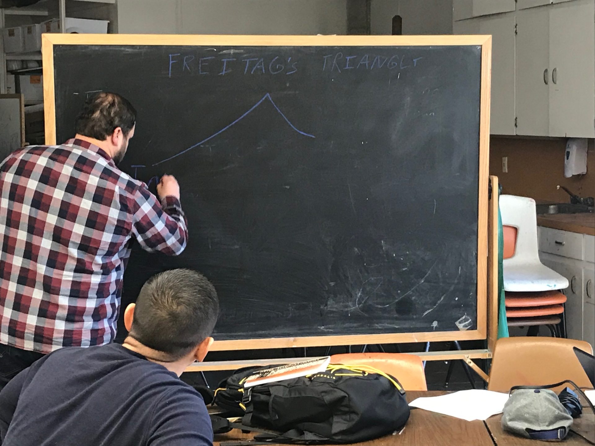 Instructor Clayton Bradshaw writing on a blackboard with a student seated at a table