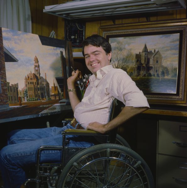 Young Randy Souders in wheelchair with paintbrush in hand beside one of his paintings