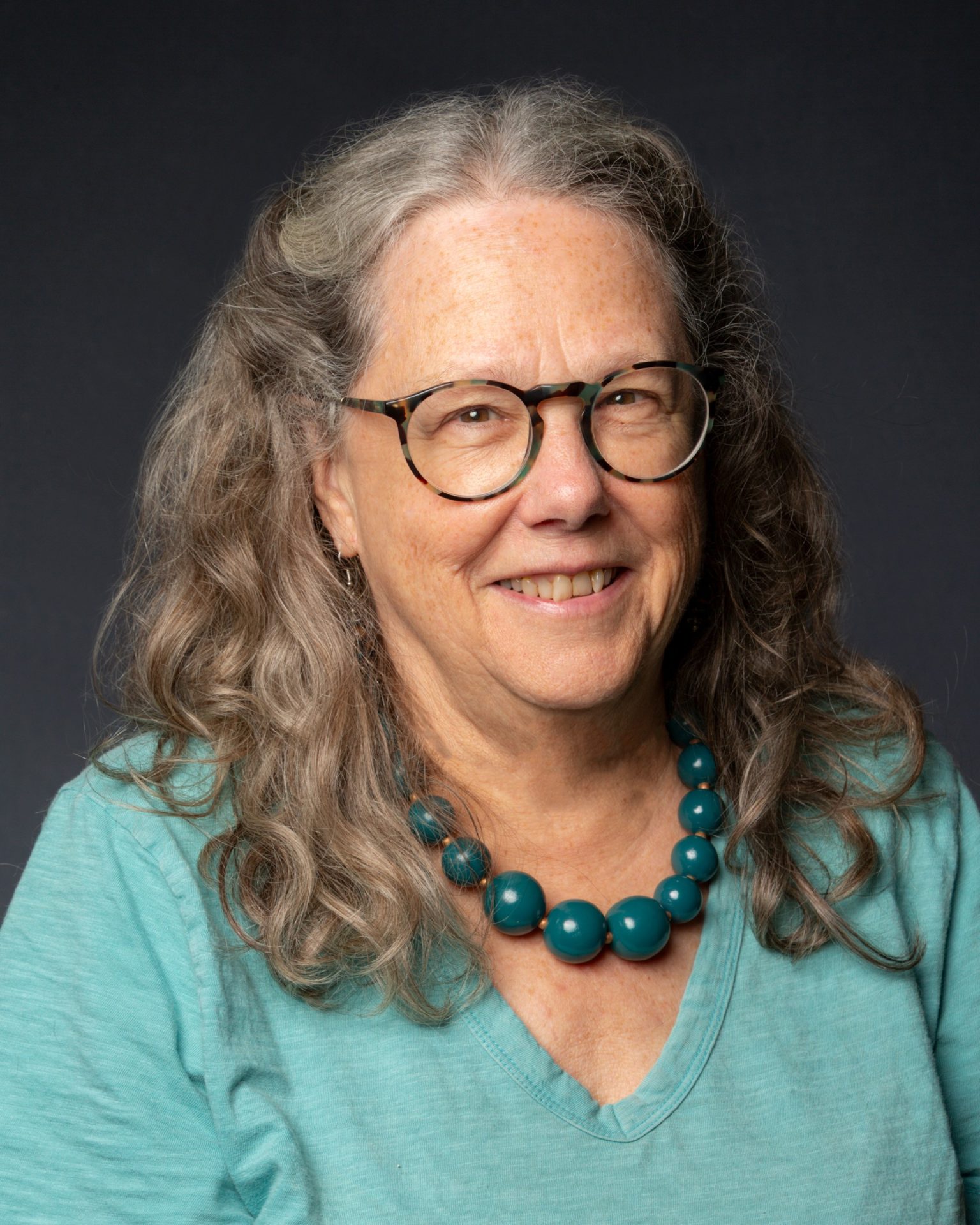 Celia Hughes wearing a light blue top with a large dark blue beaded necklace and black rimmed glasses with grey curly/wavy hair