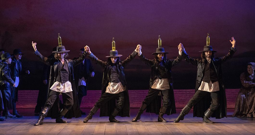 four men join hands and dance while balancing bottles on their hats