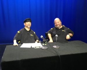 two men in wheelchairs sit behind black table with two microphones and a blue screen in the background