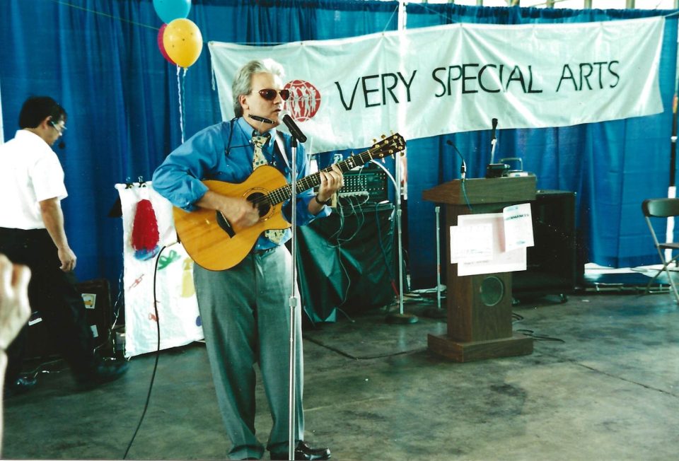 A color photo shows Jeff in his late 50s playing guitar and singing into a microphone on stage. Behind him a banner reads, “Very Special Arts.”