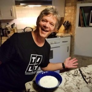 A man with short hair and a big smile strikes a pose in front of a bowl of milk in a kitchen