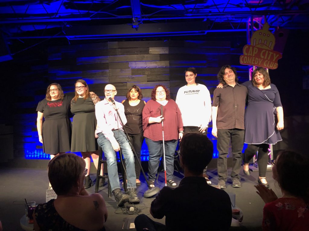 Eight comics pose for a photo on stage