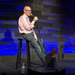 older comic with glasses sits on a stool and speaks into a microphone on stage