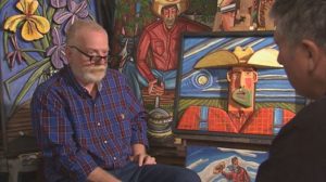 Keith Davis being interviewed with one of his cowboy paintings in the background