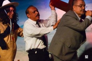 Three Latino actors walk across a stage, one carries a large wooden crucifix.