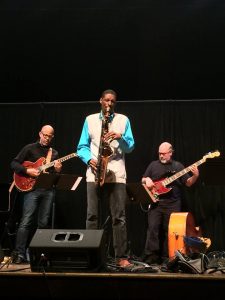 quamon playing saxophone on stage with a bass player and guitarist