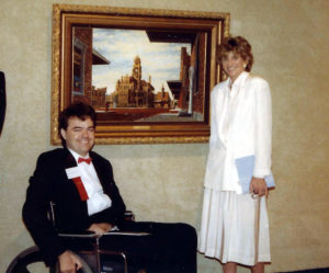 artist dressed in tuxedo and seated in wheelchair poses next to one of his paintings
